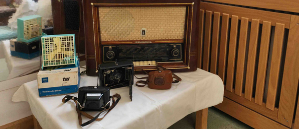 Auf einem kleinen Tisch mit einem weißen Tischtuch stehen ein altes Radio, drei alte Fotoapparate und ein Tischventilator.